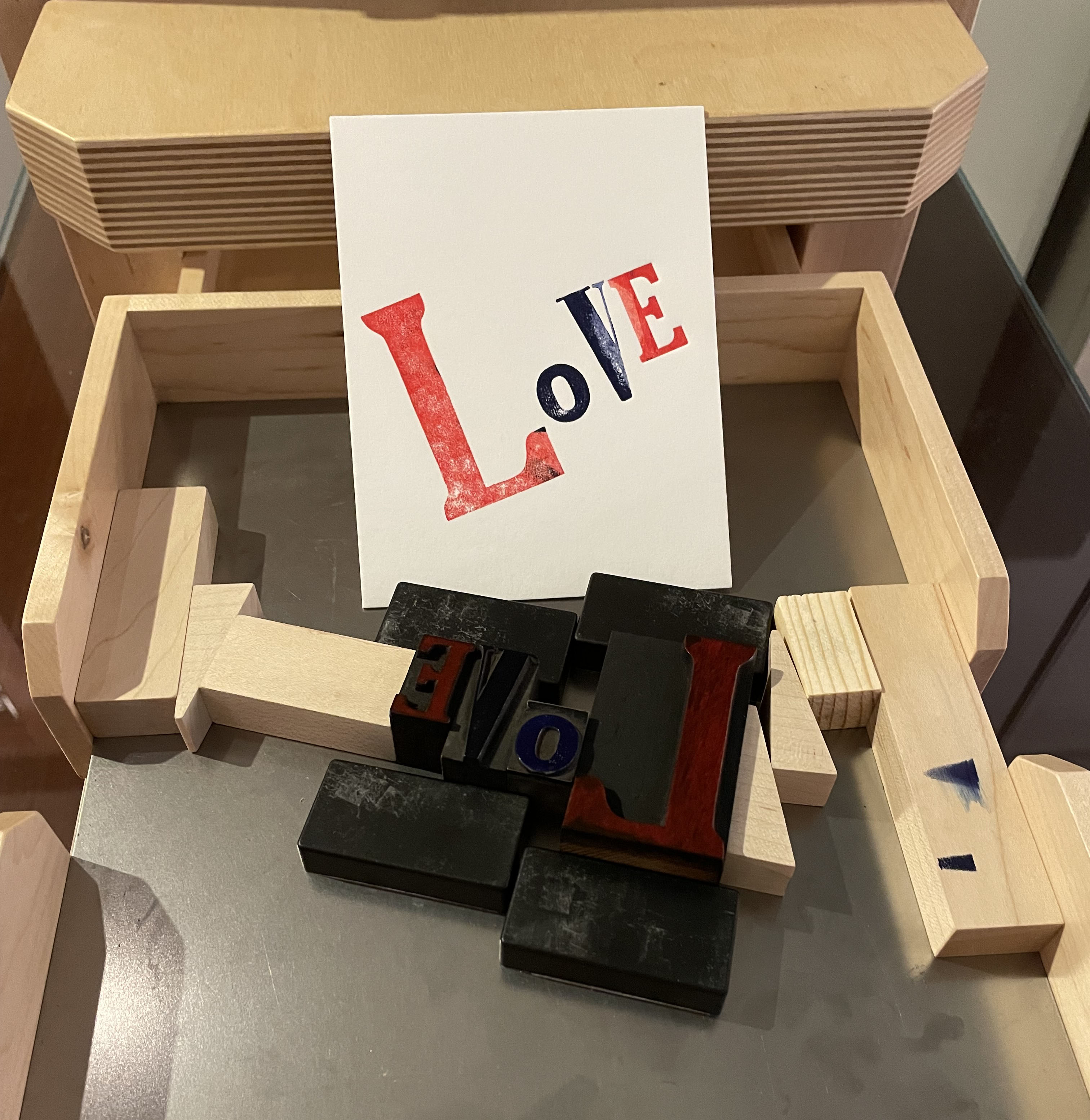 image of Book beetle tabletop press with inked wood type locked up with magnets and wood furniture spelling L-O-V-E. White notecard with print of L-O-V-E in red and blue ink