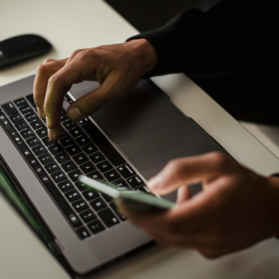 Person typing on laptop while holding a cell phone in the other hand